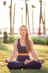 Young girl sitting in Lotus pose practicing Yoga in the park near seafront line at sunset sunrise.