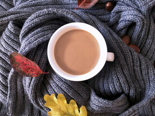 A cup of fragrant cappuccino wrapped in a gray knitted scarf decorated with bright autumn leaves and acorns.