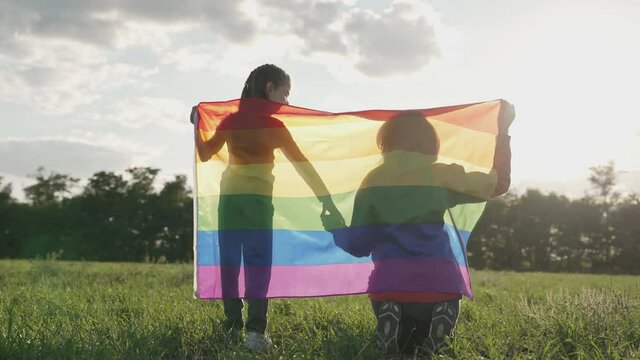 Symbol of love, freedom or LGBT pride concept. Girls holding the rainbow flag against the blue sky background, outdoors in the summer. Mom takes daughter by the hand. Liberty right