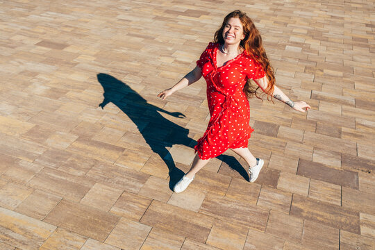 Cheerful redhead woman in red dress walking on footpath during sunny day