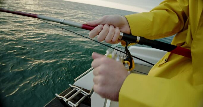 the fisherman in a yellow raincoat unwinds the fishing line while ocean fishing at sunset. Man's hands hold a fishing rod. Close up view.