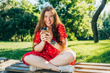 Cheerful woman holding disposable coffee cup while sitting with cross-legged on bench