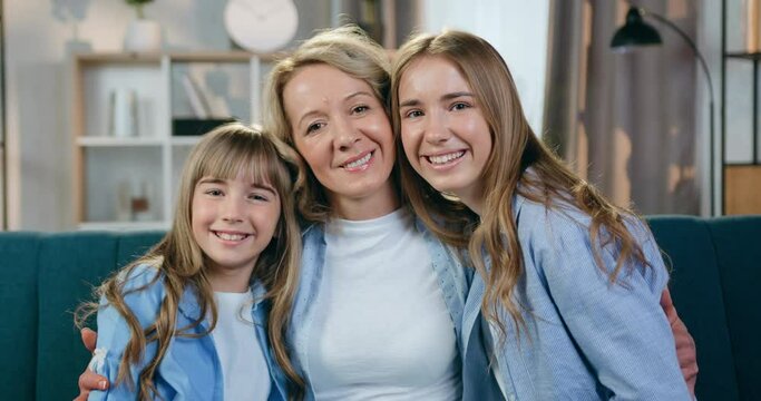 Close up portrait of lovely smiling happy blond mother and her cute friendly younger and eldest daughters which looking into camera with huggs at home