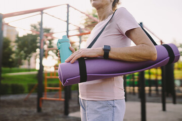 Active senior woman with fitness mat and water bottle in hands walking on workout outdoor gym....