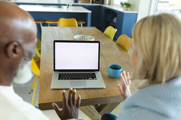 Happy senior diverse couple in kitchen sitting at table, using laptop, making video call