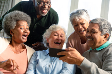 Five happy diverse senior friends sitting on sofa and looking at smartphone