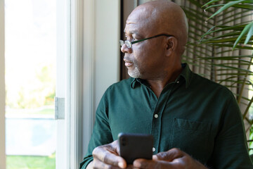 Thoughtful african american senior man holding smartphone ale looking outside window - Powered by Adobe