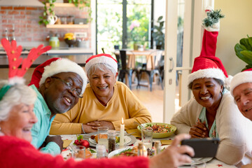 Diverse group of happy senior friends in holiday hats celebrating christmas together, taking selfie