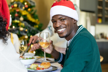 Happy african american man in santa hat toasting, celebrating christmas with friends at home
