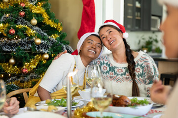 Happy diverse female friends in santa hats embracing, celebrating christmas with friends at home