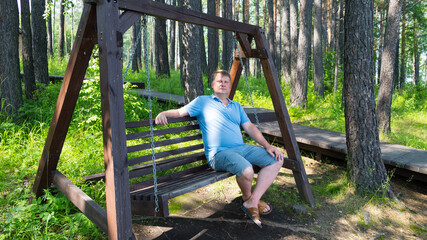 Portrait of an adult man resting in park on a wooden swing on a summer day.