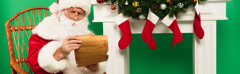 Santa claus holding paper on rocking chair near fireplace with christmas stockings on green background, banner