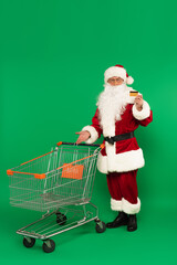 Santa claus holding credit card and pointing at shopping cart on green background