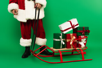 Cropped view of santa claus standing near gifts on sleigh on green background