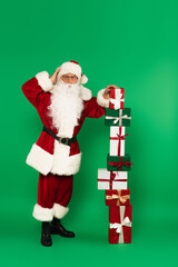 Worried santa claus standing near stacked presents on green background