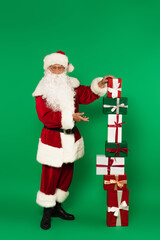Santa claus in eyeglasses and costume pointing at stacked gift boxes on green background