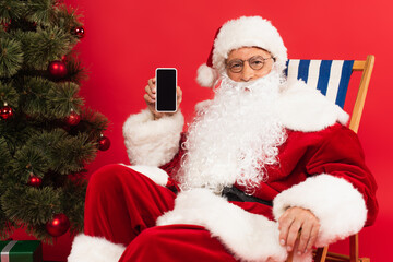 Santa claus holding smartphone on deck chair near christmas tree on red background