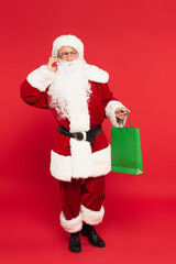 Santa claus talking on smartphone and holding shopping bag on red background