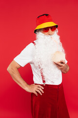 Santa claus in sunglasses and panama holding cocktail in coconut isolated on red