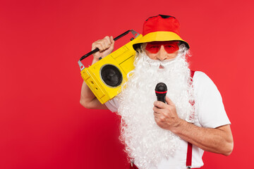 Santa claus in panama and sunglasses holding microphone and boombox isolated on red