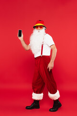 Fototapeta na wymiar Santa claus in panama and sunglasses holding smartphone on red background