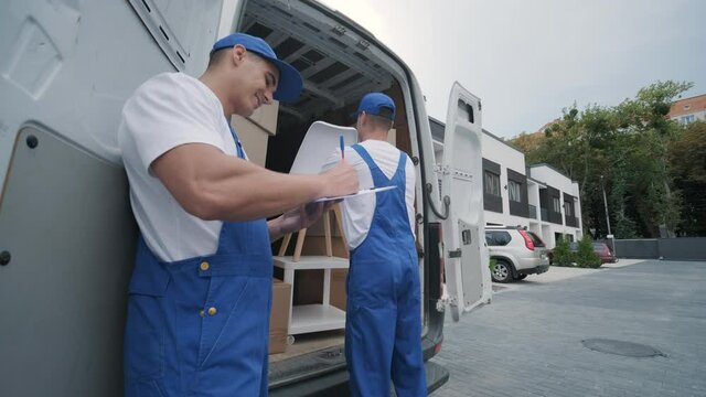 Two young workers of removal company are loading boxes and furniture into a minibus