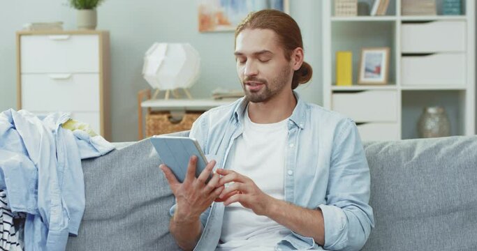Happy Caucasian man sitting at home on couch talking on video call using tablet and bragging about ticket to travel abroad.