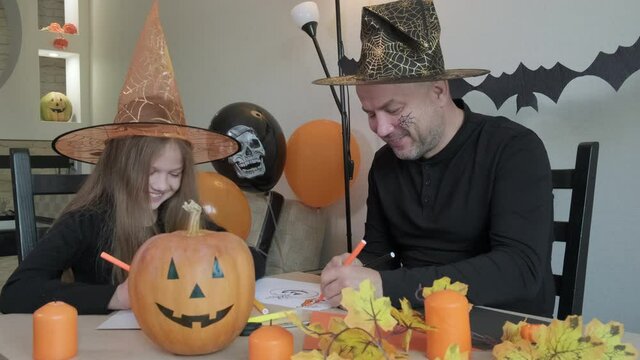 Child with father draw pictures on the theme of halloween in decorated room at home. Little girl and her dad smiling, laughing and having fun together, happy family preparing to celebrate the holiday.