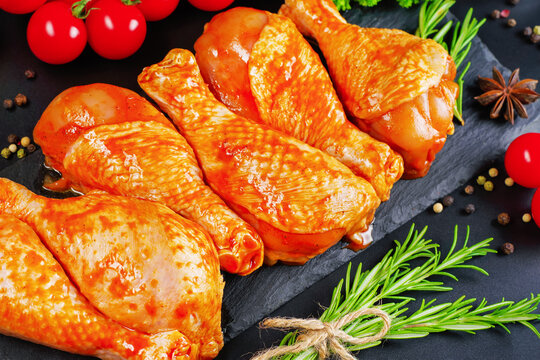Barbecue marinated chicken dramstik on a black cooking board on a black background with fresh rosemary leaves and fresh cherry tomatoes, close-up. Semi-finished product.