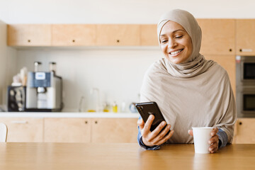 Middle eastern woman in hijab using mobile phone while drinking coffee