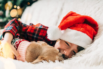 Fototapeta premium Merry Christmas. Happy little child and puppy dog in sweater playing, hug and having fun on couch with blanket at winter holidays. Kid boy in Santa hat. Christmas tree with light bokeh. Happy new year