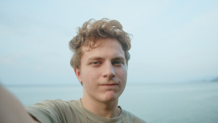 Handsome young blonde hair colored eyes man making video call chat with the phone in his hand, waving at the camera and smiling. Ocean sea background