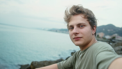 Fototapeta premium Young blond handsome boy making video call or vlogging on sea and cloudy background he pointing to the sea and smiling