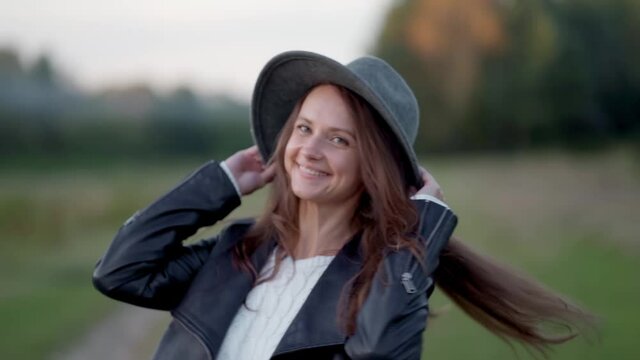 A long-haired young woman in a hat is walking alone in the park, looking at the camera, spinning, holding a hat and smiling broadly