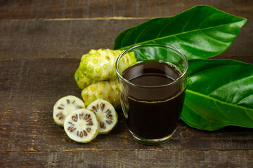 Noni juice in transparent glass and noni fruit with green leaf on wooden background. 