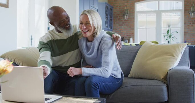 Happy senior diverse couple in living room sitting on sofa, using laptop, making video call