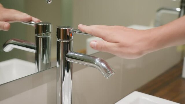 A man closes a beautiful shiny faucet against the background of a white sink and a wooden countertop. A beautiful man's hand closes the new chrome faucet, and crystal clear water stops flowing from it
