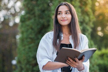 Fototapeta premium A young and beautiful Arab or Caucasian girl dressed in casual style is reading a book while sitting in the park. Knowledge and education concept.
