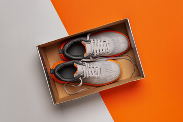 New gray textile sneakers with grooved orange sole in the open box over gray orange background....