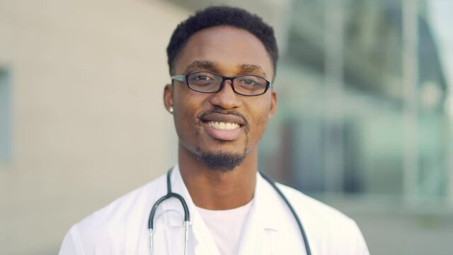 Close Up Portrait African American Doctor Looking At Camera Background Of Modern Hospital Outside. Confident Happy Medic Outdoors. Male Scientist Therapist Wearing Scrubs Near Clinic. Copy Space