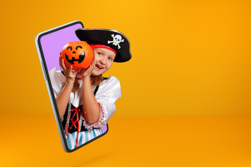 Little girl in a carnival costume of a pirate on Halloween looks from a smartphone. The child is online on the screen of a mobile phone and holds a bucket with a pumpkin lantern. Copy space.