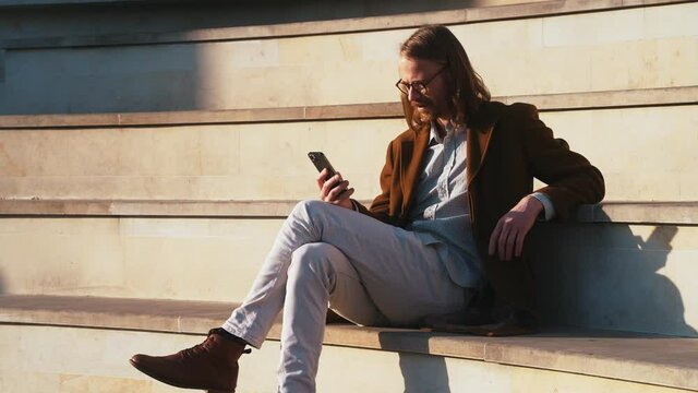 A side-view of a long-haired happy man in a coat looking at the phone while sitting on the stairs in the sun