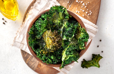 Baked kale chips in a wooden bowl on a light background. Top view. Chips from the leaves of kale cabbage close-up. Kale chips with olive oil and salt, a tasty vegan snack. - obrazy, fototapety, plakaty