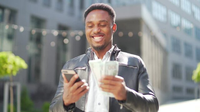 African american guy standing on street modern building rejoices show earnings winning at betting cash money with mobile phone in hands. Happy glad black man outdoors with smartphone and pack dollars
