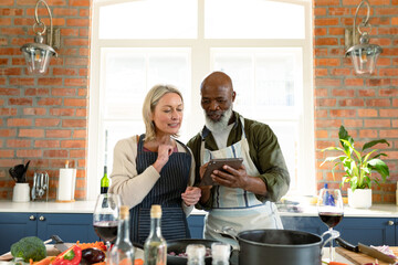 Happy senior diverse couple in kitchen wearing aprons, cooking together, using tablet