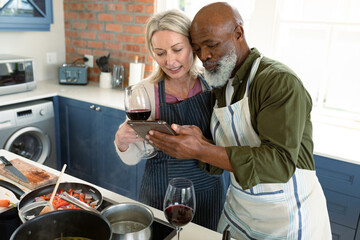 Happy senior diverse couple in kitchen wearing aprons, cooking together, using tablet