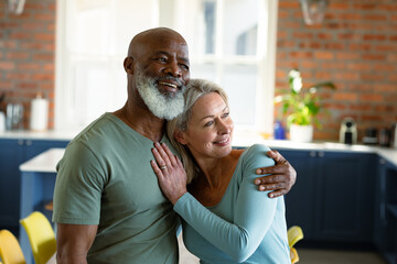 Happy senior diverse couple in kitchen embracing and smiling