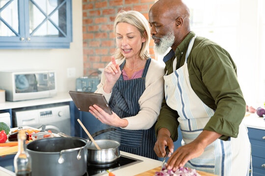 Happy senior diverse couple in kitchen wearing aprons, cooking together, using tablet - Powered by Adobe