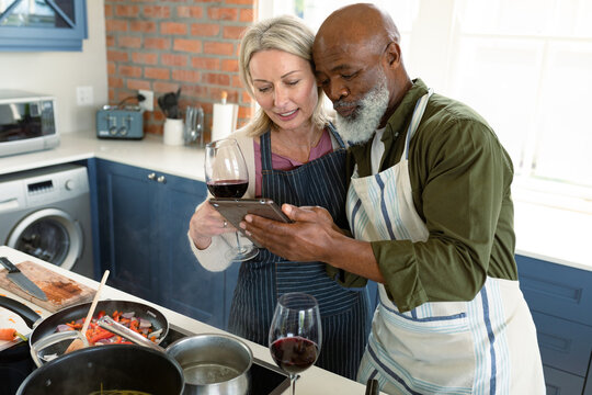 Happy senior diverse couple in kitchen wearing aprons, cooking together, using tablet
