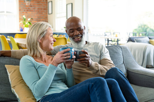 Happy senior diverse couple in living room sitting on sofa, drinking coffee